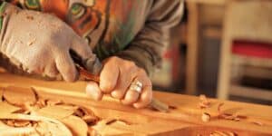 A close-up image of a person’s hands working on a piece of wood. The person is wearing a glove on the left hand and holding a chisel in the right hand, carving into the wood. Wood shavings are scattered on the work surface, indicating an active woodworking process. The background is softly blurred, with hints of furniture suggesting this activity is taking place in a workshop environment.