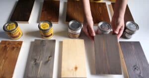 The image depicts an array of eight wooden boards on a white surface. Each board is in a different stage of varnishing, ranging from natural, untreated wood to darker shades of varnish. Four cans of varnish are aligned on the left, and four on the right. Hands are holding two cans above the boards.