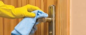 Close-up of a person’s hand wearing a yellow glove while wiping a door handle with a blue cloth, emphasizing cleanliness and hygiene in household chores.