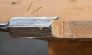 Close-up of a chisel cutting into a piece of wood. The chisel blade is partially inserted into the wood, showing layers of wood shavings and the smooth cut surface inside the incision. The background is blurred, highlighting the detail of the woodworking process.
