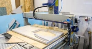 A CNC router in operation, carving a pattern into a white material in a workshop environment, surrounded by various pieces of wood and other materials.