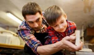 Two individuals engaged in a woodworking activity in a workshop, with one person guiding the other’s hands as they use a tool on a piece of wood. The background features various tools and wooden materials.