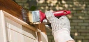 Close-up of a hand wearing a white glove with grey grip dots, holding a red-handled paintbrush applying white paint to a wooden surface, with a blurred brick wall in the background.