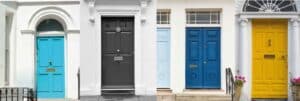 A row of four adjacent doors, each painted in a different vibrant color: turquoise, black, blue, and yellow. The doors are set within a white-walled building with architectural details like columns and decorative glass windows above them. The second door from the left has two potted plants on either side of the entrance.