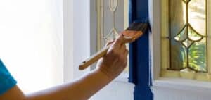 A person is painting a door frame with a brush. The paint being applied is a dark blue color. The door has decorative glass panels with a geometric design.