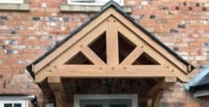 A close-up view of a wooden gable roof structure above a doorway, attached to a brick wall. The gable features a triangular design with visible wooden beams and a dark shingled roof.