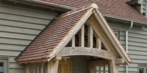 Wooden porch with a gabled roof and reddish-brown shingles on a house with greenish-gray siding.