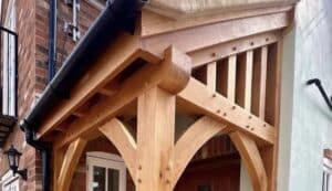 A close-up view of a wooden porch attached to a brick house. The porch features intricate woodwork with multiple beams and joints, and a black gutter system above.