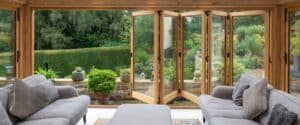 A cozy living room with large bi-fold wooden doors open to a lush green garden. The room features two gray sofas with cushions and a central ottoman. Outside, there are various potted plants and a well-maintained hedge, with a dense forest in the background.