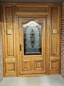 A wooden front door with a decorative glass panel in the centre. The door is surrounded by wooden panels and brick walls on either side. The glass panel features an intricate design with frosted and clear sections, and a metal handle is on the left side of the door.