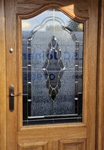 A wooden door with a decorative glass panel in the centre. The glass panel features an intricate design with frosted and clear sections, framed by metal outlines. The door has a brass handle and a keyhole below it.