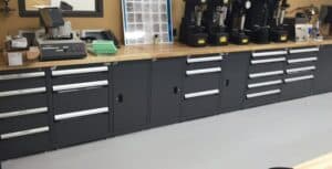 Well-organized laboratory workspace with a long wooden countertop and multiple black storage cabinets underneath. On the countertop, there are various pieces of laboratory equipment, including a Struers machine, several hardness testers, a chart displaying different material samples, boxes, papers, and green plastic trays. The setup highlights the tools and organization necessary for conducting precise and accurate tests, relevant for fields such as materials science, engineering, or quality control.