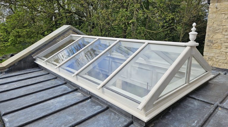 A rooftop skylight with multiple glass panels set in a white frame. The skylight is installed on a sloped roof with a dark, ribbed surface. Trees and greenery are visible in the background.